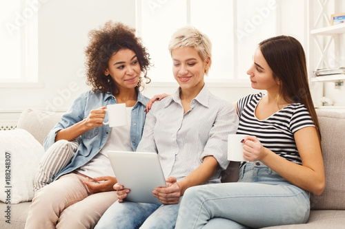 Three happy female friends using tablet and drinking coffee