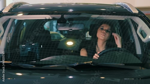 Brunette Business Successful Woman in Luxury Car salon holding hands on steering wheel. She in black dress. Car is black.