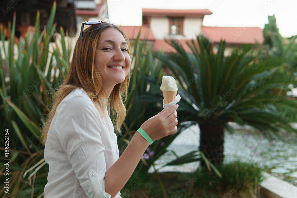 Fototapeta premium Portrait of beautiful positive young woman eating ice cream on background of palm trees.