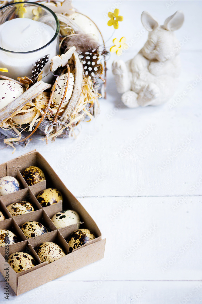 Easter spring decorative composition, crafted wreath with candle inside, white bunny and box with quail eggs. Close up portrait on white wooden background with place for text. Easter spring decorative composition, crafted wreath with candle inside, white bunny and box with quail eggs. Close up portrait on white wooden background with place for text.