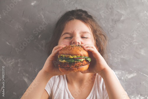 The little girl is eating a healthy baked sweet potato burger with a whole grains bun, guacamole, vegan mayonnaise and vegetables. Child vegan concept, gray background.