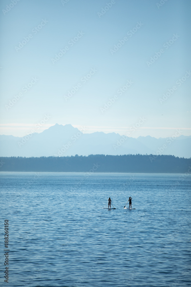 Two silhouetted stand up paddle board riders on a calm blue ocean, with mountains on the horizon