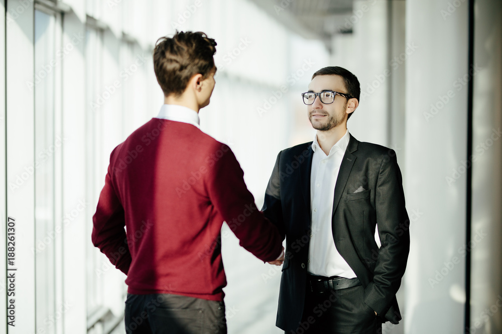 Two smiling businessmen shaking hands together while standing by windows in an office boardroom overlooking the city Two smiling businessmen shaking hands together while standing by windows in an office boardroom overlooking the city