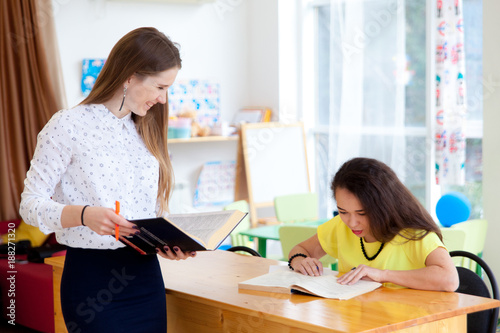 Young woman teacher helps the female student to understand a lesson. Student sits at her desk with a large book in the classroom and listening to teacher.