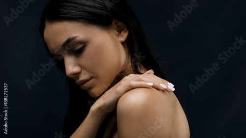 Side view of a beautiful woman with black hair touching her shoulder with hand against black background