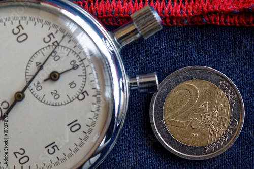 Euro coin with a denomination of 2 euro and stopwatch on worn blue jeans with red stripe backdrop - business background