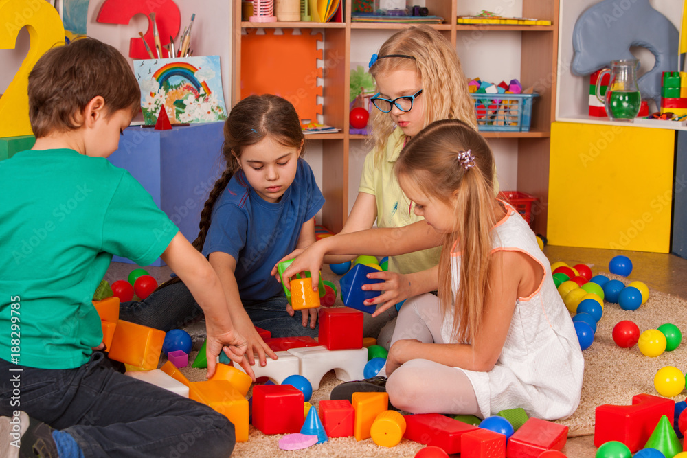 Fototapeta premium Children building blocks in kindergarten. Group kids playing toy on floor. Top view of interior preschool. Building a tower of cubes. Newcomer in children's team.