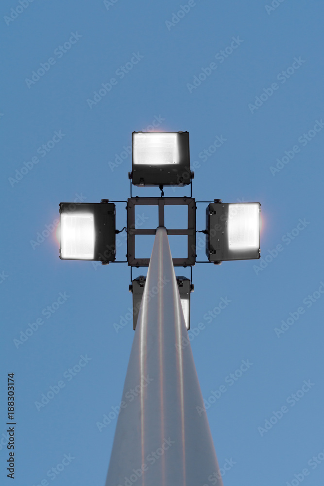 street lamp with four floodlights shines with yellow light in the evening against a blue dark sky, perspective view from the bottom of a round post street lamp with four floodlights shines with yellow light in the evening against a blue dark sky, perspective view from the bottom of a round post
