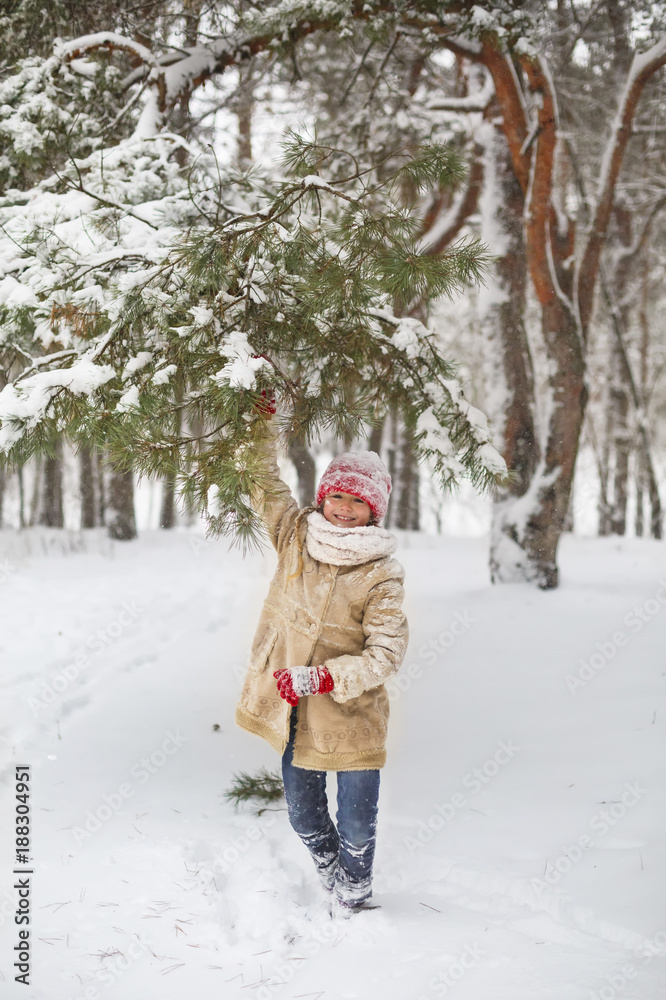 Beautiful little girl have fun in snowy winter forest. Adorable child wearing a red knitted hat and jacket walk outdoor at Christmas. Kid playing in snowy forest. Family winter vacation in mountains