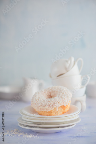 White donut with coconut topping on a light background with porcelain cups and saucers. High key food photography. Stack of tableware.