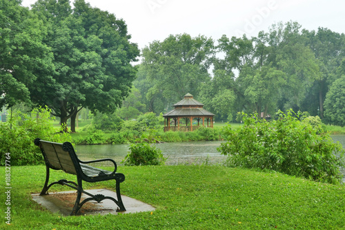 Beautiful summer rainy day nature background. Summer landscape in a city park with a bench on a foreground and wooden gazebo on a background during warm rainy day.