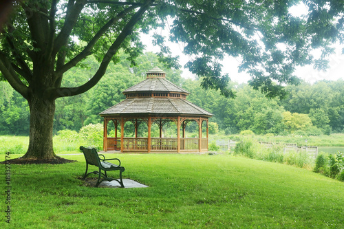 Fototapeta Naklejka Na Ścianę i Meble -  Beautiful summer rainy day nature background. Summer landscape in a city park with a bench on a foreground and wooden gazebo on a background during warm rainy day.