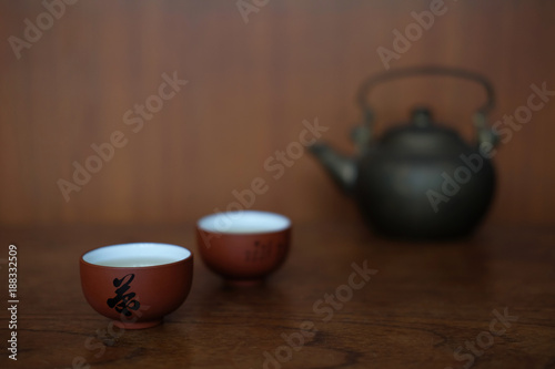 CHINESE TEA CUP WITH BLACL IRON KETTLE Small ceramic Chinese tea cups are placed together with black iron kettle on the wood table in front of wood wall.