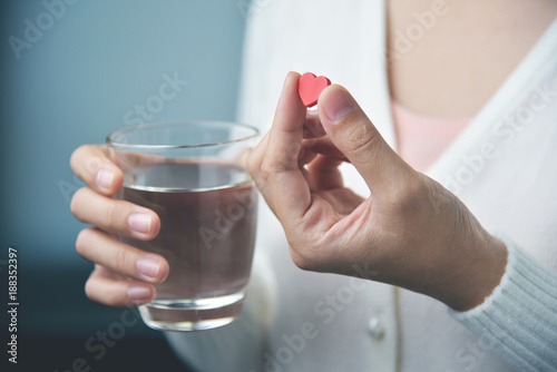 Female hands with white shirt is holding red pill in heart shape and glass of water