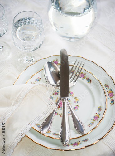 Elegant dinner set with silver knife, fork and spoon lying across luxyry porcelain dishes, and crystal glassware on cream vintage lace tablecloth photo