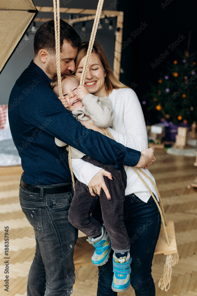 Mom and dad play with charming little son on a swing in the room - obrazy, fototapety, plakaty 