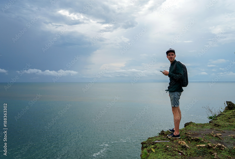 A young man with a backpack, standing on a cliff and pointing his hand to the distance. He sets a goal.