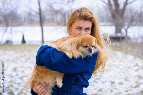Woman pet owner with blue coat and golden hair walking during winter with her dog. Young lady with pekingese dog walking on the snowy field. Tender feeling with a dog 