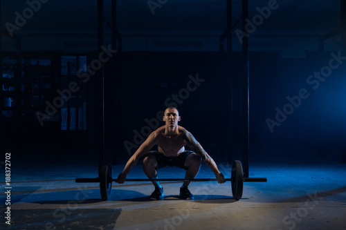 A sporty man lifts a bar in the gym in a dark light