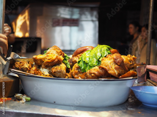 Thai local traditional street food stall, fat pork leg boiled in black sticky gravy, piled with green kale, on big white aluminum bowl on dirty selling trolley, traffic people and car, night market 