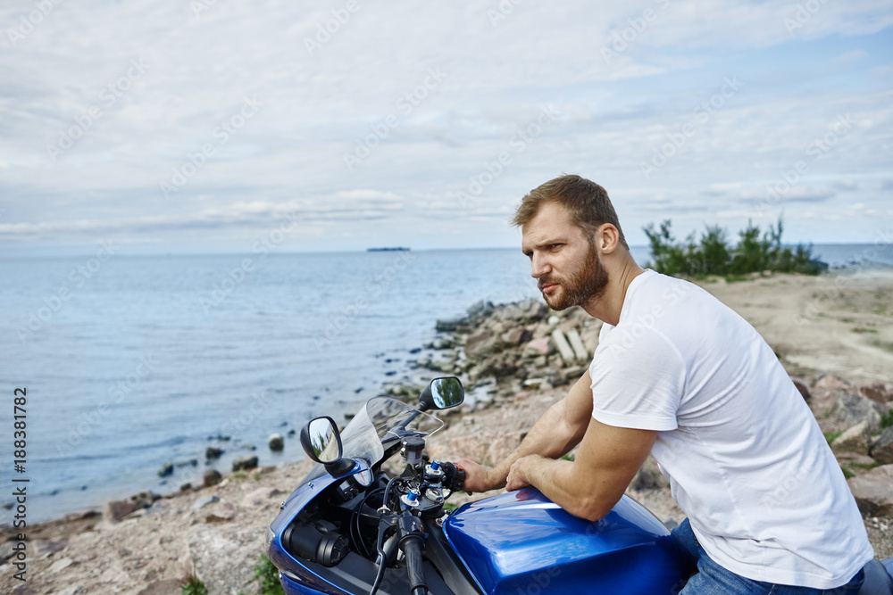 People, nature, travel and freedom concept. Picture of handsome young Caucasian male wearing white t-shirt relaxing by the sea, leaning on his blue motorbike and looking around, enjoying view