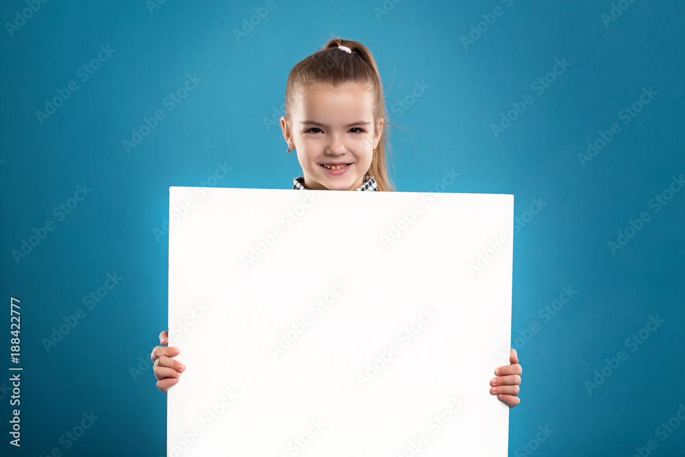 Isolated on blue, beautiful caucasian brunette child hold empty poster, look at camera, stand behind the poster Isolated on blue, beautiful caucasian brunette child hold empty poster, look at camera, stand behind the poster