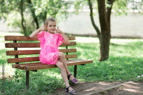 Little caucasian girl sitting in park on bench. A child in a beautiful pink dress in the open air