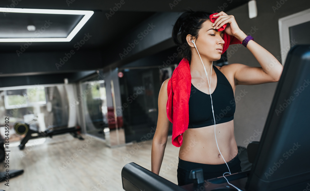 Sporty young woman athlete running on treadmill in the gym, wiped off with a red towel and listening the music by her earphones. Sport, fitness, healthy life and people concept. Sporty young woman athlete running on treadmill in the gym, wiped off with a red towel and listening the music by her earphones. Sport, fitness, healthy life and people concept.