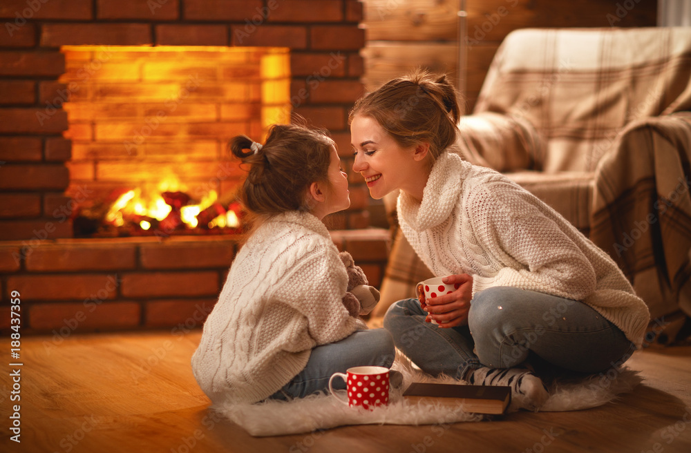 family mother and child drinking tea and laughing on winter evening by fireplace. - obrazy, fototapety, plakaty 