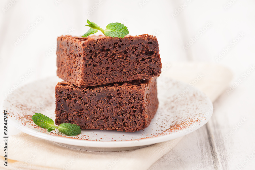 Chocolate brownie square pieces in stack on white plate decorated with mint leaves and cocoa powder on white vintage wooden background. American traditional delicious dessert. Close up photography Chocolate brownie square pieces in stack on white plate decorated with mint leaves and cocoa powder on white vintage wooden background. American traditional delicious dessert. Close up photography