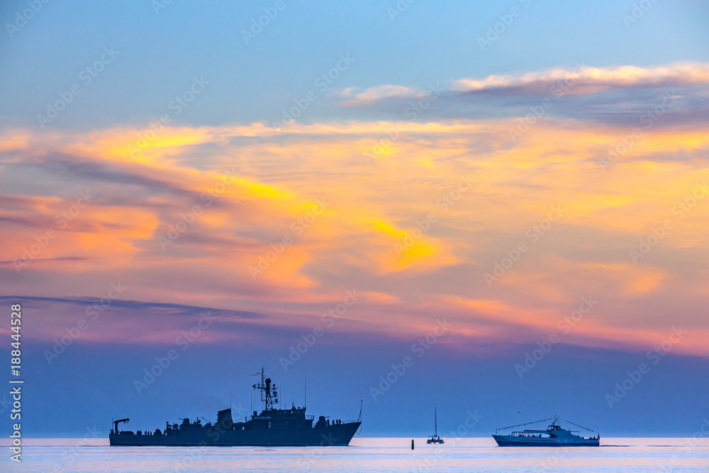 Crossroads on the sea, the silhouette of a warship in front of a boat and a yacht in the light of the sunset under the yellow beautiful clouds. Coastal seascape on the Black Sea, Sochi, Russia.