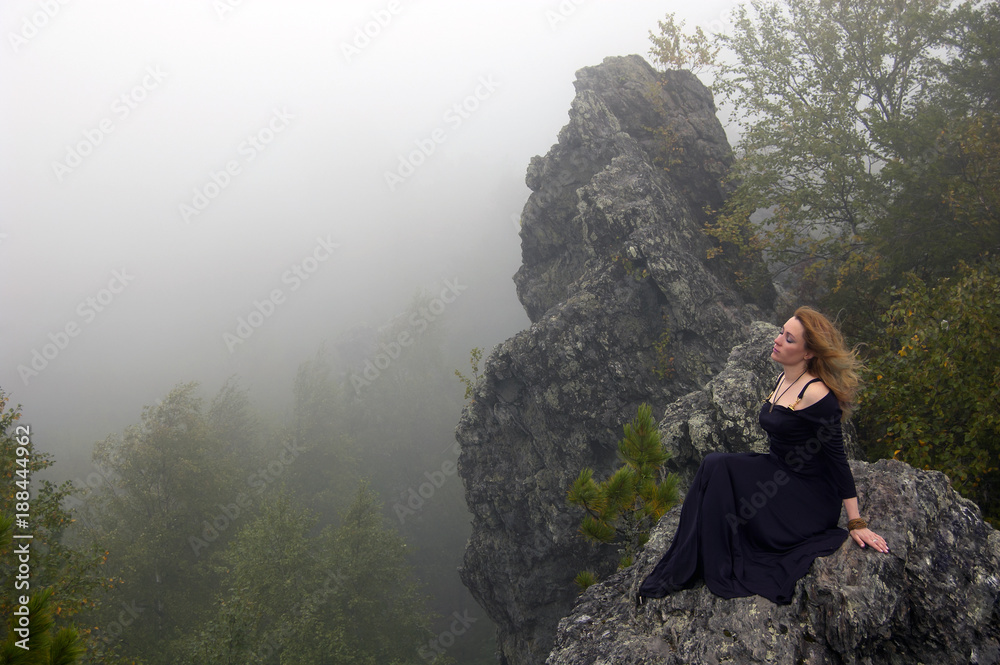 beautiful young woman in a black dress with red hair flying by the wind, sits on a cliff towering above the forest in the fog