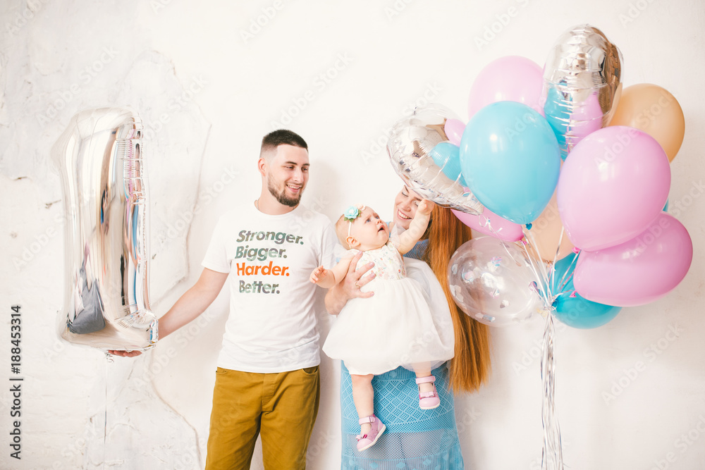 young family, parents mom and dad holding a baby girl one year on a white background at home. The concept of a children's family holiday is decorated with inflatable airy multi-colored balls