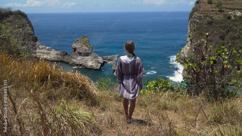 Young girl stands on the edge of a cliff and looks at the sea. Girl on the edge of the cliff enjoys the view of the ocean. Atuh beach on Nusa Penida island. Travel concept.