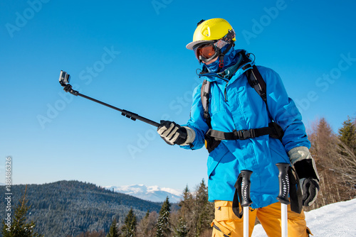 Shot of a skier in winter clothing taking a selfie using monopod for his camera. Blue sky and winter forest on the background skiing activity sports concept