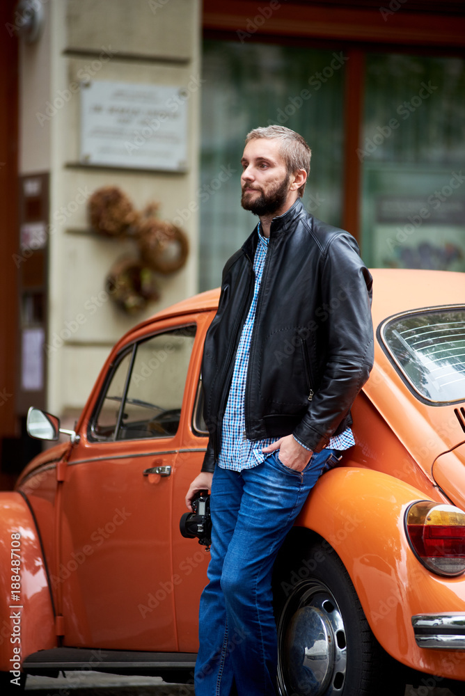 Profile of a man with the beard leaning on orange retro car and holding in hand a professional photo camera against the backdrop of urban architecture in Budapest. Close-up