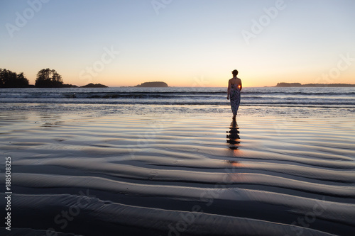 Woman Walking on a Sandy Beach during a vibrant and colorful summer sunset. Taken in Tofino  Vancouver Island  BC  Canada.