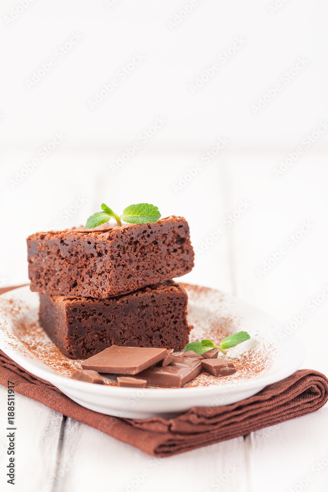 Chocolate brownie square pieces in stack on white plate decorated with mint leaves and cocoa powder on white vintage wooden background. American traditional delicious dessert. Close up photography Chocolate brownie square pieces in stack on white plate decorated with mint leaves and cocoa powder on white vintage wooden background. American traditional delicious dessert. Close up photography