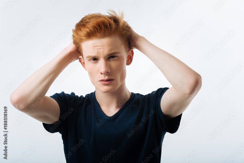 Earnest. Handsome unsmiling red-haired young man touching his head with his hands and wearing a black t-shirt and looking seriously Earnest. Handsome unsmiling red-haired young man touching his head with his hands and wearing a black t-shirt and looking seriously