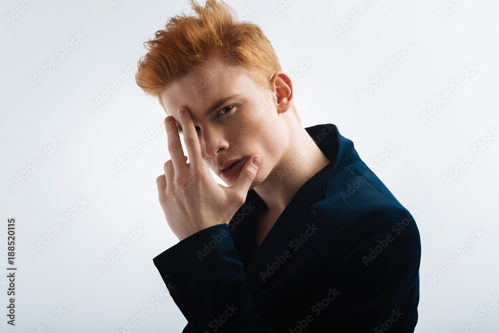 Fashion plate. Good-looking humourless red-headed young man wearing a black jacket and touching his forehead and staring Fashion plate. Good-looking humourless red-headed young man wearing a black jacket and touching his forehead and staring
