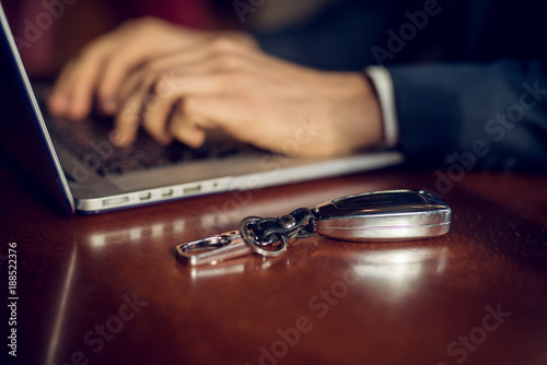 Close up focus view of car keys on the desk while hands of young successful stylish businessman in the suit typing on a laptop in a cafe or restaurant.