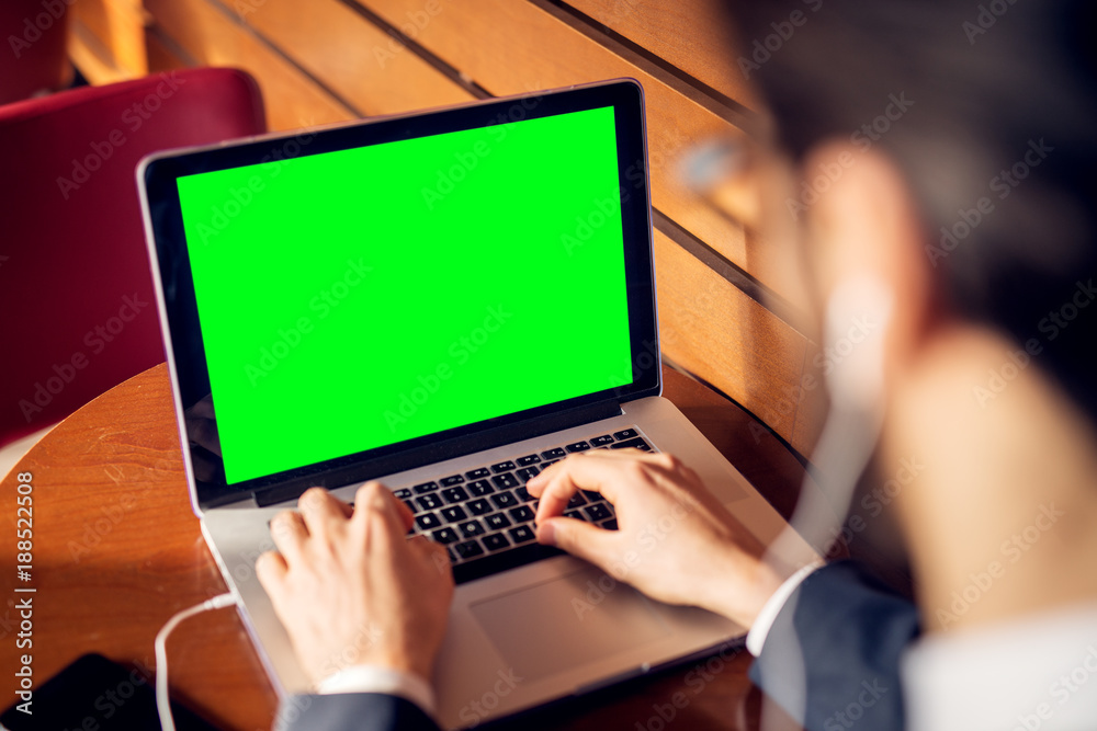Rear close up focus view laptop with blank green screen and hands of young successful stylish focused businessman in the suit with earphones typing in a cafe or restaurant near the wooden wall. Rear close up focus view laptop with blank green screen and hands of young successful stylish focused businessman in the suit with earphones typing in a cafe or restaurant near the wooden wall.