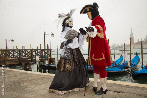 Venice, Italy - February 22, 2017: Venice Carnival - Couple in love of Venetian masks on St. Mark's Square in Venice with Gondolas in background.