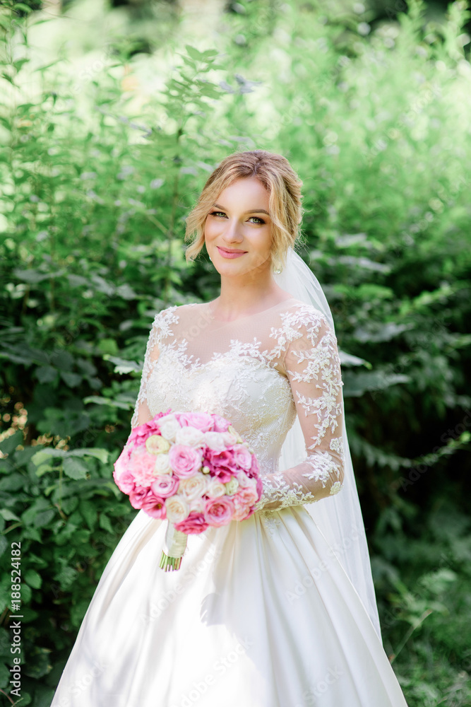 Bride in classy dress with long sleeves holds pink wedding bouquet standing in the park Bride in classy dress with long sleeves holds pink wedding bouquet standing in the park