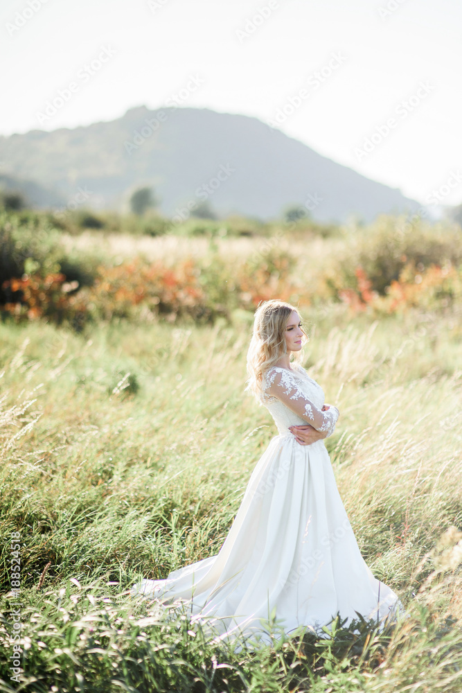 Stunning bride with white curls stands on the field in a sunny day