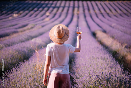 Young woman drink rose wine in the sunset lavender field, standing back to the camera, Provence, south France