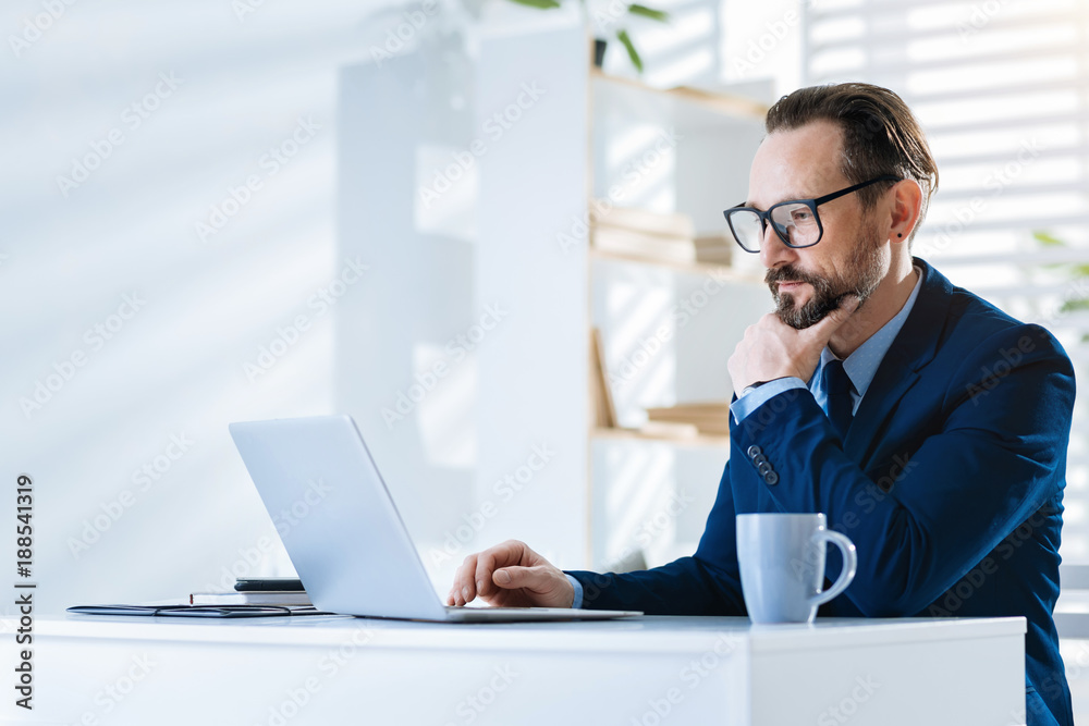 Working process. Thoughtful pleasant attractive man touching chin while using laptop and wearing glasses Working process. Thoughtful pleasant attractive man touching chin while using laptop and wearing glasses