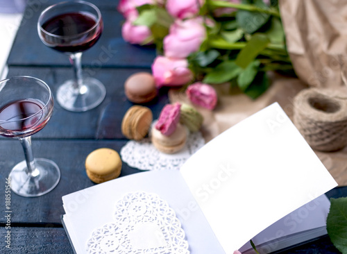 A bouquet of roses is pink, in gray paper and a white heart. Open notebook. Two glasses of red wine. Sweet pasta macaroons of different colors, on Valentine's Day. A free place for text or advertising