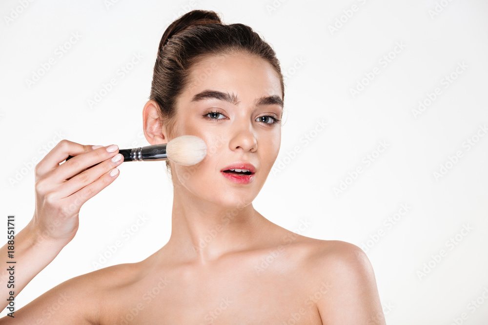 Close up image of tender half-naked woman with healthy skin applying powder with soft brush and looking at camera isolated over white background