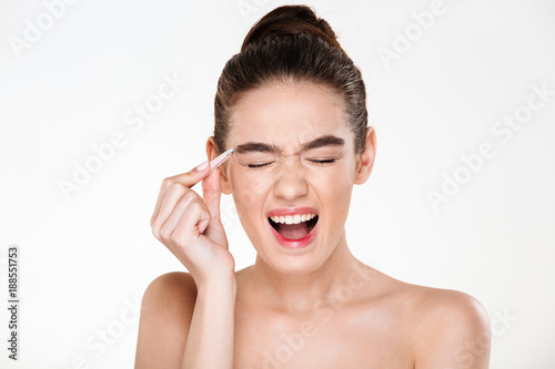 Beauty portrait of sensual brunette woman with hair in bun screaming in pain while plucking eyebrows with tweezers isolated over white background photo